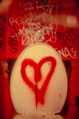 A bathroom stall features a toilet with a white seat, upon which a large red heart is spray-painted. The background shows walls covered with various pieces of graffiti in different colors and styles.