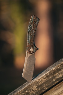 An intricately designed knife stands vertically on a wooden surface. The handle features a textured pattern with a mix of dark and metallic hues. The blade is thick and appears robust, casting a shadow on the surface below. The background is blurred, consisting of warm, earthy tones that give a natural ambiance.