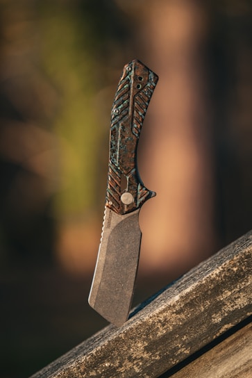 An intricately designed knife stands vertically on a wooden surface. The handle features a textured pattern with a mix of dark and metallic hues. The blade is thick and appears robust, casting a shadow on the surface below. The background is blurred, consisting of warm, earthy tones that give a natural ambiance.