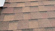 A close-up view of asphalt roof shingles arranged in a uniform pattern. The shingles have a mix of brown, red, and gray tones, with a textured, granular surface. The edges are aligned, showing a layered installation.