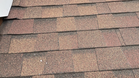 A close-up view of asphalt roof shingles arranged in a uniform pattern. The shingles have a mix of brown, red, and gray tones, with a textured, granular surface. The edges are aligned, showing a layered installation.