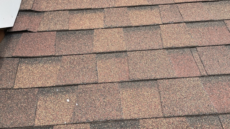 A close-up view of asphalt roof shingles arranged in a uniform pattern. The shingles have a mix of brown, red, and gray tones, with a textured, granular surface. The edges are aligned, showing a layered installation.