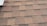 A close-up view of asphalt roof shingles arranged in a uniform pattern. The shingles have a mix of brown, red, and gray tones, with a textured, granular surface. The edges are aligned, showing a layered installation.