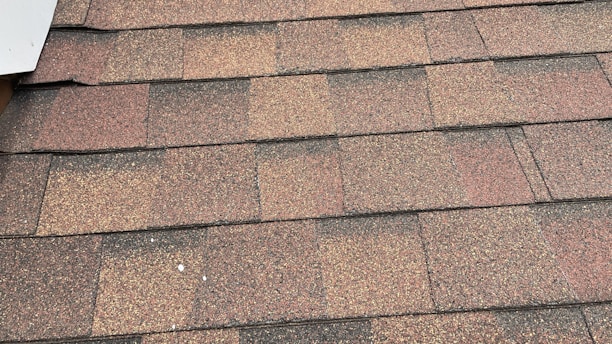 A close-up view of asphalt roof shingles arranged in a uniform pattern. The shingles have a mix of brown, red, and gray tones, with a textured, granular surface. The edges are aligned, showing a layered installation.