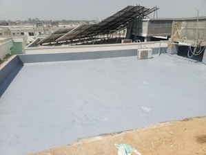 A flat rooftop with a smooth, gray surface, featuring a solar panel array set up at an angle. An air conditioning unit is visible on the roof, and there are ropes and wires hanging along the side. The background shows a cityscape under a clear sky.