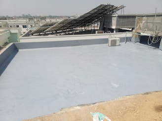 A flat rooftop with a smooth, gray surface, featuring a solar panel array set up at an angle. An air conditioning unit is visible on the roof, and there are ropes and wires hanging along the side. The background shows a cityscape under a clear sky.