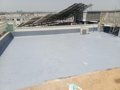 A flat rooftop with a smooth, gray surface, featuring a solar panel array set up at an angle. An air conditioning unit is visible on the roof, and there are ropes and wires hanging along the side. The background shows a cityscape under a clear sky.