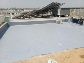 A flat rooftop with a smooth, gray surface, featuring a solar panel array set up at an angle. An air conditioning unit is visible on the roof, and there are ropes and wires hanging along the side. The background shows a cityscape under a clear sky.
