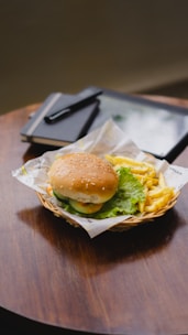 A hamburger with lettuce, tomato, and a sesame seed bun is served on a basket lined with paper next to a portion of crispy golden fries. The basket is placed on a round wooden table. In the background, there's a closed notebook and a pen on top of a tablet.