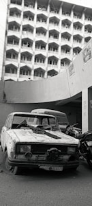 An abandoned, weathered car is positioned in front of a large building with numerous identical windows. The car appears damaged and neglected, with a broken windshield and license plate visible. Motorcycles are parked beside the car, and the building in the background is multi-storied and bears signs of urban decay.