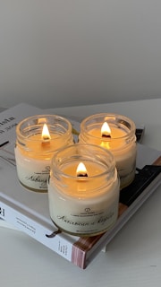 Three lit candles in glass jars are placed atop a closed book. The candles have a soft, warm glow which casts a cozy ambiance. The jars are labeled with text, suggesting different scents. The book cover is partially visible, adding a touch of sophistication to the scene.