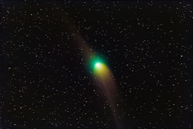 A comet with a bright green glow at the head and a long, trailing tail is surrounded by a vast expanse of stars in the night sky.