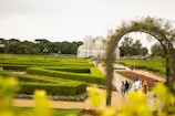 A lush, meticulously landscaped garden with geometric hedges and pathways. A striking glass greenhouse, with an elegant dome structure, is visible in the background. Groups of people are walking along the paths, enjoying the serene surroundings. An arch made of plants and flowers adds a charming touch to the scene.
