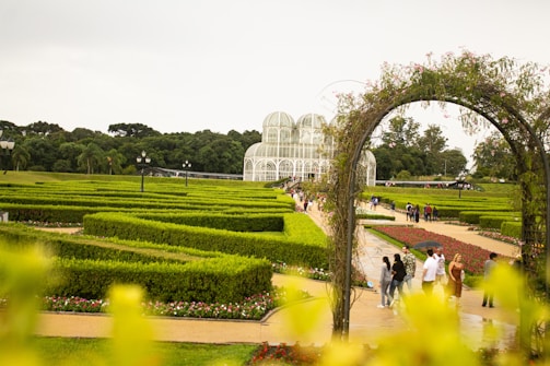 A lush, meticulously landscaped garden with geometric hedges and pathways. A striking glass greenhouse, with an elegant dome structure, is visible in the background. Groups of people are walking along the paths, enjoying the serene surroundings. An arch made of plants and flowers adds a charming touch to the scene.