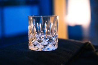A clear, intricately cut glass tumbler sits on a dark surface. The background features soft lighting with a blue and warm tone, creating a cozy ambiance.