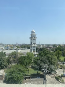 A tall, white minaret stands prominently in the center of a landscape. It has a large dome on top, with several smaller balconies attached to the structure. The foreground features a lush green area with numerous trees, and buildings are visible in the background under a clear blue sky.