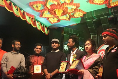 A group of individuals stands together indoors, some holding plaques or awards. The ceiling is decorated with colorful drapery in red and yellow hues. The people are dressed in winter clothing and formal attire, such as sweaters and suits. The atmosphere seems festive or ceremonial.