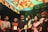 A group of individuals stands together indoors, some holding plaques or awards. The ceiling is decorated with colorful drapery in red and yellow hues. The people are dressed in winter clothing and formal attire, such as sweaters and suits. The atmosphere seems festive or ceremonial.