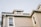 A light brown dog is peering out from a small attic window on the roof of a beige house with white trims. The house has multiple windows and a slightly tilted sloping roof.