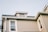 A light brown dog is peering out from a small attic window on the roof of a beige house with white trims. The house has multiple windows and a slightly tilted sloping roof.