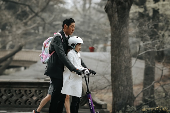 A person dressed in a suit is assisting a child who is wearing a helmet and riding a scooter. The scene is set in a park with bare trees and a stone bridge, giving it an autumnal or early spring feel.