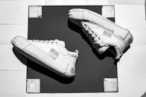 A pair of white sneakers with black laces are arranged on a black square mat. The sneakers have distinctive rope-like patterns around the soles and tags with text attached to the sides. The background consists of a white surface with a grid-like pattern and worn edges.