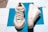 Two off-white sneakers are placed on a bright blue background. One shoe is positioned upright, showcasing the lace-up design, while the other rests on its side, revealing the sole and side details. The sneakers feature textured soles and visible stitching, with inscriptions on the shoe’s tongue and front.