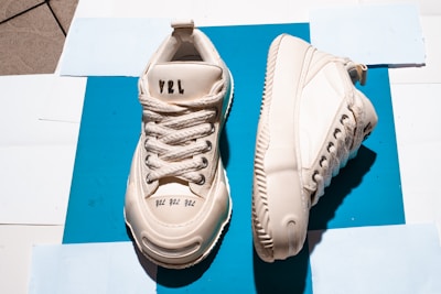 Two off-white sneakers are placed on a bright blue background. One shoe is positioned upright, showcasing the lace-up design, while the other rests on its side, revealing the sole and side details. The sneakers feature textured soles and visible stitching, with inscriptions on the shoe’s tongue and front.
