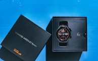 A smart watch is displayed in its box against a blue background. The box is black and labeled 'HUAWEI WATCH GT Runner' with a mention of 'Powered by HarmonyOS'. The watch has a round face with a black band and features several details in orange and white.