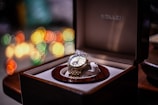 A luxury wristwatch with a gold band and intricate design nestled in a wooden box with a velvet lining. The background features a bokeh effect with colorful, circular lights in shades of yellow, orange, green, and red, adding a festive and elegant feel.
