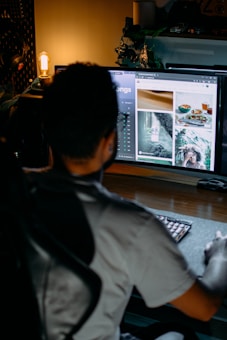 A person is seated at a desk in a dimly lit room, facing a large computer monitor displaying images and text. A warm light bulb is glowing in the background, and there are plants and decorative items surrounding the monitor. The scene creates a cozy and focused atmosphere.