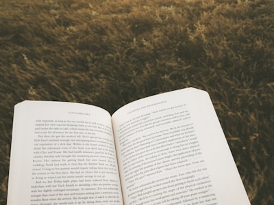 An open book is held against a backdrop of grass or field, suggesting a peaceful or serene outdoor reading setting.
