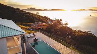An elevated view of a serene landscape featuring a partially shadowed villa with a swimming pool and deck chairs. Rolling hills covered in dense greenery stretch along the coast. The setting sun casts a golden light across a calm body of water in the background, creating a peaceful and tranquil atmosphere.