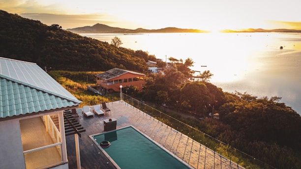 An elevated view of a serene landscape featuring a partially shadowed villa with a swimming pool and deck chairs. Rolling hills covered in dense greenery stretch along the coast. The setting sun casts a golden light across a calm body of water in the background, creating a peaceful and tranquil atmosphere.
