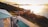 An elevated view of a serene landscape featuring a partially shadowed villa with a swimming pool and deck chairs. Rolling hills covered in dense greenery stretch along the coast. The setting sun casts a golden light across a calm body of water in the background, creating a peaceful and tranquil atmosphere.