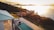 An elevated view of a serene landscape featuring a partially shadowed villa with a swimming pool and deck chairs. Rolling hills covered in dense greenery stretch along the coast. The setting sun casts a golden light across a calm body of water in the background, creating a peaceful and tranquil atmosphere.