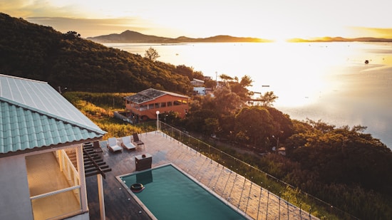 An elevated view of a serene landscape featuring a partially shadowed villa with a swimming pool and deck chairs. Rolling hills covered in dense greenery stretch along the coast. The setting sun casts a golden light across a calm body of water in the background, creating a peaceful and tranquil atmosphere.