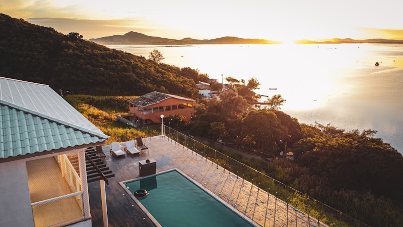An elevated view of a serene landscape featuring a partially shadowed villa with a swimming pool and deck chairs. Rolling hills covered in dense greenery stretch along the coast. The setting sun casts a golden light across a calm body of water in the background, creating a peaceful and tranquil atmosphere.