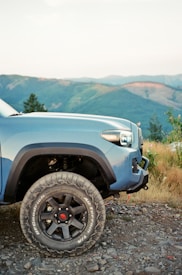 A rugged blue off-road vehicle is parked on a rocky terrain with expansive mountain views in the background. The vehicle has notable large off-road tires, and the area is surrounded by grassy vegetation and small shrubs.