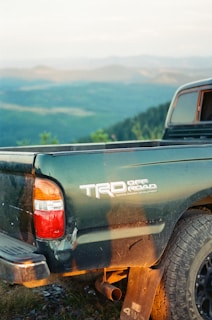 A green TRD Off-Road Toyota truck is parked on a rocky terrain with expansive mountains and forests visible in the background. The tailgate and rear tire of the truck are prominently visible.