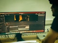 A computer monitor displays video editing software with a timeline and preview of a scene. The scene shows two silhouettes against a warm, orange backdrop. A person is seated at a desk beside the monitor, with computer equipment, including a keyboard and a Mac Mini, visible on the desk.