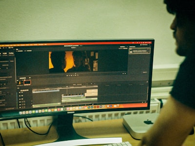 A computer monitor displays video editing software with a timeline and preview of a scene. The scene shows two silhouettes against a warm, orange backdrop. A person is seated at a desk beside the monitor, with computer equipment, including a keyboard and a Mac Mini, visible on the desk.