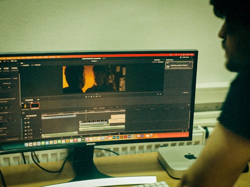 A computer monitor displays video editing software with a timeline and preview of a scene. The scene shows two silhouettes against a warm, orange backdrop. A person is seated at a desk beside the monitor, with computer equipment, including a keyboard and a Mac Mini, visible on the desk.