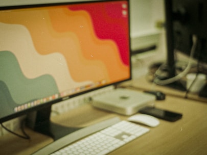 A desktop setup featuring a large monitor with a colorful, wavy pattern as the wallpaper. In front of the monitor, there is a white keyboard and a white mouse. To the side of the monitor, there is a small silver computer, likely a mini-PC. The background includes another monitor that is partially visible and several cables on the desk.