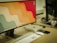 A desktop setup featuring a large monitor with a colorful, wavy pattern as the wallpaper. In front of the monitor, there is a white keyboard and a white mouse. To the side of the monitor, there is a small silver computer, likely a mini-PC. The background includes another monitor that is partially visible and several cables on the desk.