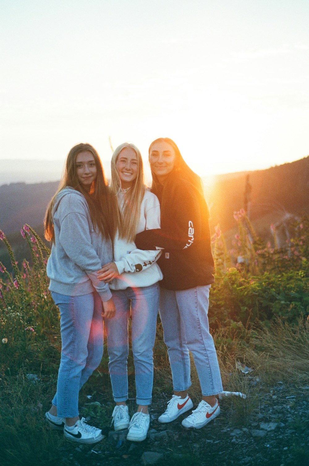 três meninas estão de pé em uma colina ao pôr do sol