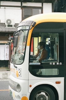 A bus is parked on a street with a driver seated inside wearing a mask. The bus exterior is mostly white with orange accents, and there is street signage and buildings visible in the background.