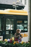 An elderly person wearing a brown hat and face mask stands near a yellow and white bus with Japanese characters. The person is holding a bag and appears to be waiting or boarding. The foreground has out-of-focus colorful flowers, while the background features part of a building and street lamps.