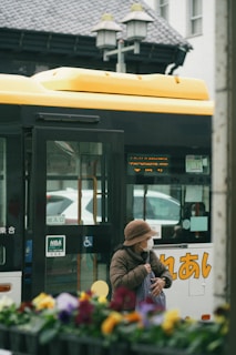 An elderly person wearing a brown hat and face mask stands near a yellow and white bus with Japanese characters. The person is holding a bag and appears to be waiting or boarding. The foreground has out-of-focus colorful flowers, while the background features part of a building and street lamps.