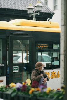 An elderly person wearing a brown hat and face mask stands near a yellow and white bus with Japanese characters. The person is holding a bag and appears to be waiting or boarding. The foreground has out-of-focus colorful flowers, while the background features part of a building and street lamps.